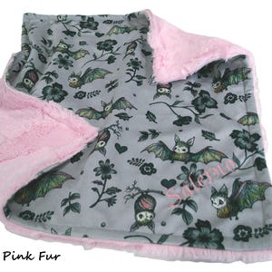 May include: A soft blanket featuring a gray fabric side with a repeating pattern of bats, flowers, and hearts, and a plush blush pink fur side. The name Salerno is embroidered in pink thread on the gray side of the blanket.