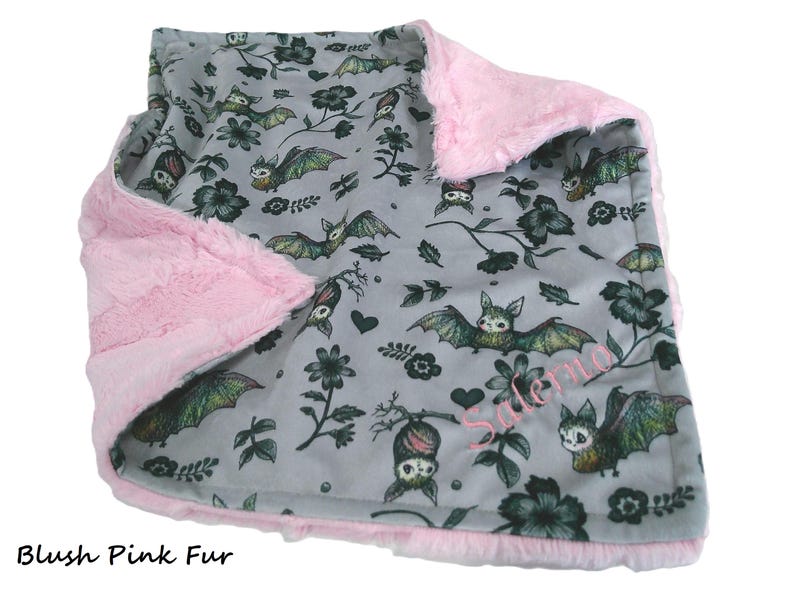 May include: A soft blanket featuring a gray fabric side with a repeating pattern of bats, flowers, and hearts, and a plush blush pink fur side. The name Salerno is embroidered in pink thread on the gray side of the blanket.
