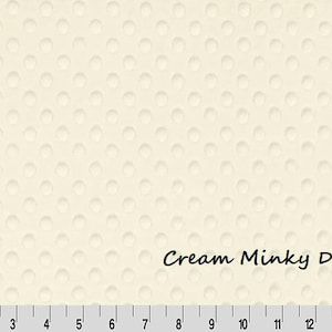 May include: Cream-colored Minky Dot fabric with a textured surface of raised dots. The text "Cream Minky Dot" is printed on the fabric. A ruler is at the bottom, with markings in inches.