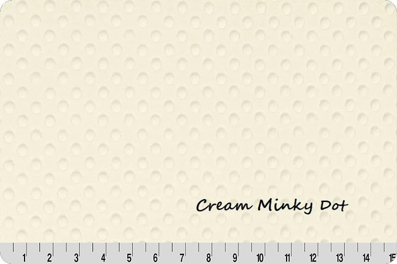May include: Cream-colored Minky Dot fabric with a textured surface of raised dots. The text "Cream Minky Dot" is printed on the fabric. A ruler is at the bottom, with markings in inches.