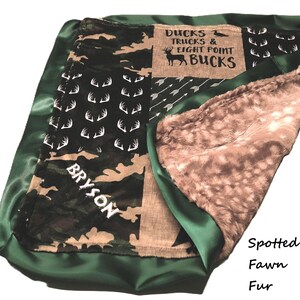 May include: A baby blanket with a green satin border and a camouflage and deer antler pattern. The blanket has a brown and white spotted fawn fur interior. Text on the blanket reads "Ducks Trucks & Eight Point Bucks" and "Bryson".