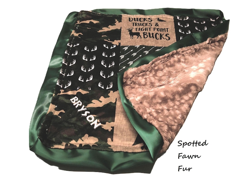 May include: A baby blanket with a green satin border and a camouflage and deer antler pattern. The blanket has a brown and white spotted fawn fur interior. Text on the blanket reads "Ducks Trucks & Eight Point Bucks" and "Bryson".