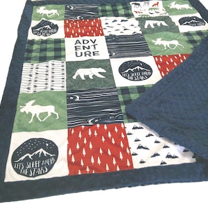 May include: A patchwork quilt featuring forest and mountain-themed designs. The quilt has a navy blue border and a navy blue textured backing. The squares display images of moose, bears, trees, and the text "ADVENTURE" and "LET'S SLEEP UNDER THE STARS".