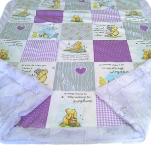 May include: A patchwork quilt featuring Winnie the Pooh illustrations, purple gingham squares, and gray wood-grain patterns. The fabric includes text such as 'It never hurts to keep looking for sunshine' and 'Sometimes the smallest things take up the most room in your heart.' The quilt is bordered by a textured light purple plush fabric.