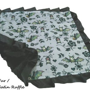 May include: A square blanket featuring a light gray fabric with a repeating pattern of green and brown bats, black flowers, and small hearts. The blanket is bordered by a wide, gathered black satin ruffle trim. Text reads: Black Fur / Black Satin Ruffle.