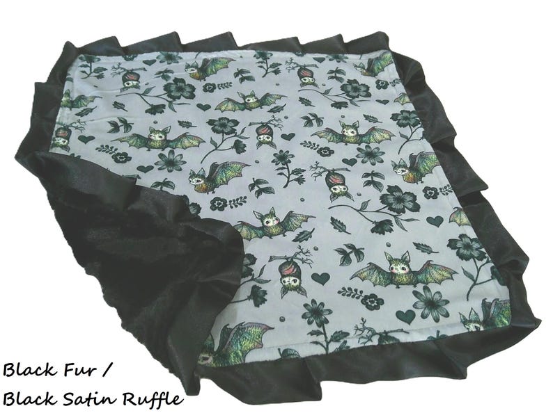 May include: A square blanket featuring a light gray fabric with a repeating pattern of green and brown bats, black flowers, and small hearts. The blanket is bordered by a wide, gathered black satin ruffle trim. Text reads: Black Fur / Black Satin Ruffle.