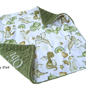 May include: A white baby blanket with a pattern of green and tan dragons, rainbows, and stars. The border is olive green minky dot fabric. The name "Ogi" is embroidered in white on the corner.