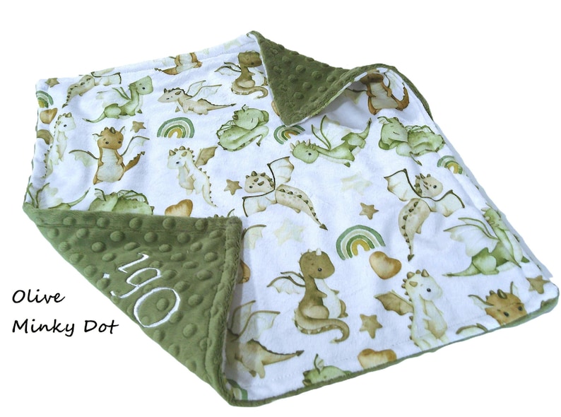 May include: A white baby blanket with a pattern of green and tan dragons, rainbows, and stars. The border is olive green minky dot fabric. The name "Ogi" is embroidered in white on the corner.