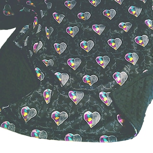 Pu&ograve; includere: Una coperta scura con un motivo di cuori a met&agrave; riempiti con contorni bianchi e disegni geometrici colorati. La coperta ha un lato inferiore verde scuro strutturato e un motivo ripetuto di cuori e viti.