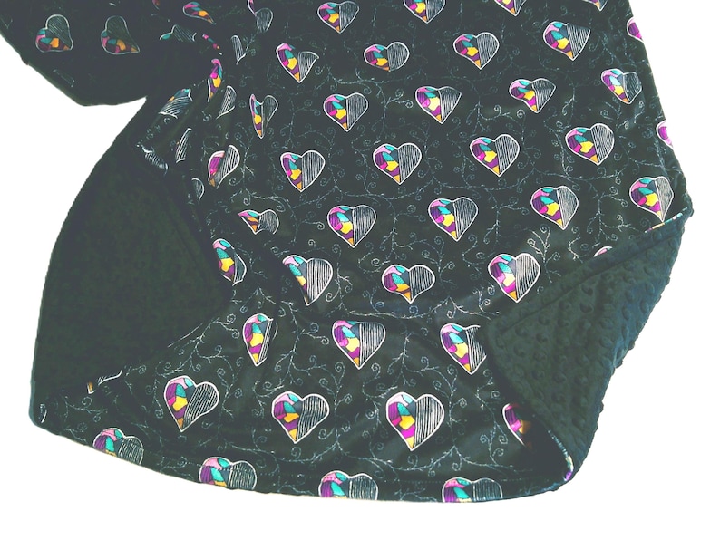 Pu&ograve; includere: Una coperta scura con un motivo di cuori a met&agrave; riempiti con contorni bianchi e disegni geometrici colorati. La coperta ha un lato inferiore verde scuro strutturato e un motivo ripetuto di cuori e viti.