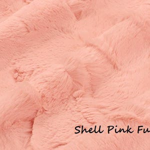 May include: Close-up of a soft, shell pink faux fur fabric. The plush material has a wavy texture with long, flowing fibers. The words "Shell Pink Fur" are written in a black script font at the bottom of the image.