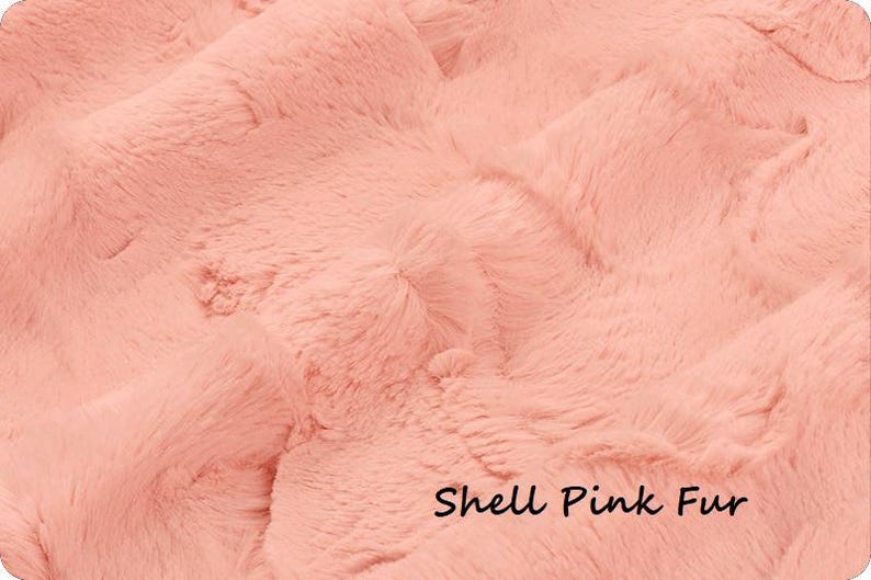 May include: Close-up of a soft, shell pink faux fur fabric. The plush material has a wavy texture with long, flowing fibers. The words "Shell Pink Fur" are written in a black script font at the bottom of the image.