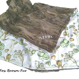 May include: A baby blanket with a brown fur top and a white bottom. The white bottom has a pattern of green and tan dragons, rainbows, and stars. The blanket has the word "AZRIEL" embroidered on the brown fur.