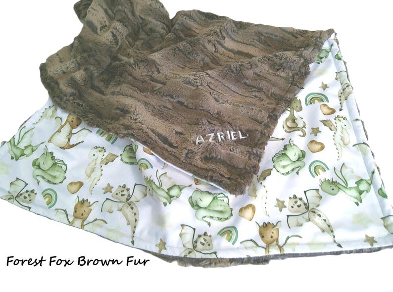 May include: A baby blanket with a brown fur top and a white bottom. The white bottom has a pattern of green and tan dragons, rainbows, and stars. The blanket has the word "AZRIEL" embroidered on the brown fur.