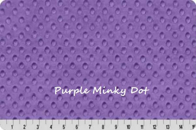 May include: A close-up of a purple minky dot fabric with a textured surface. The fabric has a repeating pattern of raised dots. The text "Purple Minky Dot" is written across the fabric. A ruler is at the bottom.