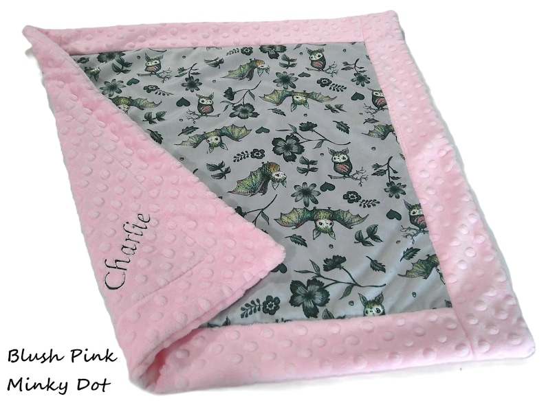 May include: A rectangular baby blanket featuring a light gray center panel with a repeating pattern of owls, bats, and dark flowers. The blanket has a textured blush pink minky dot border, with the name Charlie embroidered in black script.
