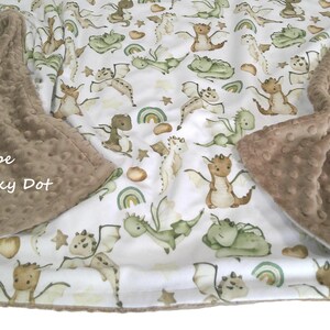 May include: A baby blanket with a white background featuring a pattern of cartoon dragons in shades of green, brown, and tan. The blanket has a taupe minky dot border. The text "Taupe Minky Dot" is visible.