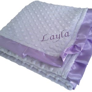 May include: A soft, white baby blanket with a textured, dotted surface. The blanket has a lavender satin trim and the name "Layla" embroidered in lavender thread. The blanket is folded, showing the layers of fabric.