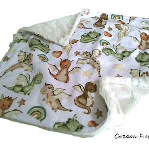 May include: A baby blanket with a white background and a pattern of cartoon dragons in shades of brown, green, and tan. The blanket has a cream-colored, plush fur trim. The text "Cream Fur" is in the bottom right corner.