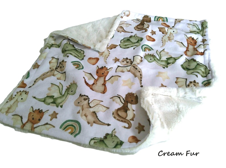 May include: A baby blanket with a white background and a pattern of cartoon dragons in shades of brown, green, and tan. The blanket has a cream-colored, plush fur trim. The text "Cream Fur" is in the bottom right corner.