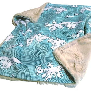 May include: A baby blanket with a blue and white wave pattern on one side and a soft, beige faux fur on the other. The blanket is rectangular and has a plush, cozy appearance, perfect for snuggling.