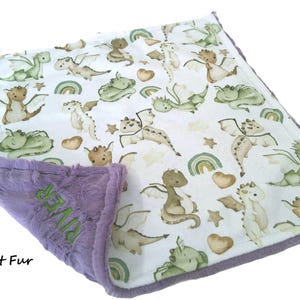 May include: A baby blanket with a white background featuring a pattern of green, brown, and tan dragons, rainbows, stars, and hearts. The border is purple, with the text "RIVER" embroidered in green. The bottom edge reads "Violet Fur".