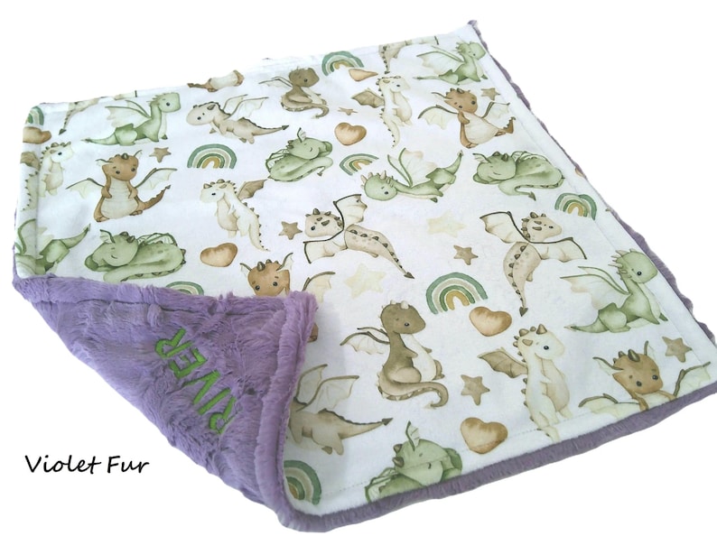 May include: A baby blanket with a white background featuring a pattern of green, brown, and tan dragons, rainbows, stars, and hearts. The border is purple, with the text "RIVER" embroidered in green. The bottom edge reads "Violet Fur".
