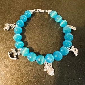 May include: A blue gemstone bracelet with silver charms, including a rocking horse, a baby bottle, a teddy bear, and a baby onesie with the word "BABY" on it.