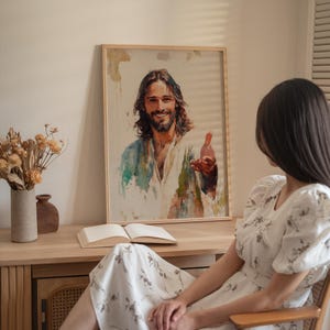 May include: A framed watercolor painting of a smiling man with long brown hair and a beard, wearing a white and blue robe. The painting is displayed on a wooden table with a book and vase.