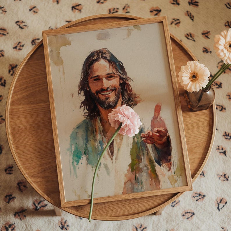 May include: A framed watercolor print of a smiling man with long hair and a beard, wearing a white and blue robe. A pink flower is in the foreground. The artwork is displayed on a wooden table with a vase of flowers.