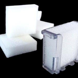 May include: A clear plastic filter housing filled with white foam, next to several white foam cubes. The items are set against a black background. The filter housing has a rectangular shape with a clear plastic frame.