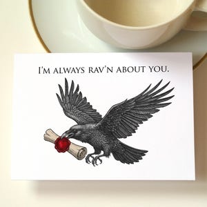 May include: Greeting card featuring a black raven illustration holding a scroll sealed with red wax. The text "I'M ALWAYS RAV'N ABOUT YOU" is printed above the raven. A white teacup and saucer are in the background.