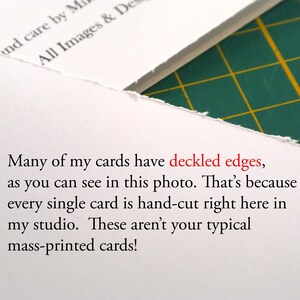 Op de afbeelding: Een witte kaart met een ruwe, onregelmatige geschepte rand. De kaart bevat zwarte gedrukte tekst die luidt: Many of my cards have deckled edges, as you can see in this photo. That&rsquo;s because every single card is hand-cut right here in my studio.