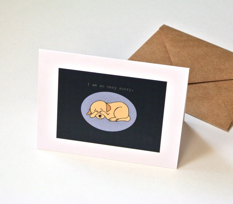 Op de afbeelding: Een witte wenskaart met een donker rechthoekig middenstuk met een lichtpaarse ovaal waarin een gele cartoonhond zijn ogen bedekt met zijn poten. Boven de hond staat de tekst, I am so very sorry. Een bruine envelop is achter de kaart geplaatst.