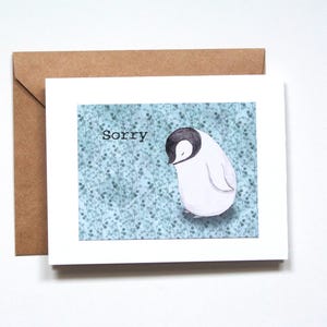 Peut inclure: Une carte de vœux blanche avec le mot "Sorry" et une illustration à l'aquarelle d'un pingouin. Le pingouin a une tête noire et se détache sur un fond bleu avec un motif floral. Une enveloppe marron est derrière la carte.