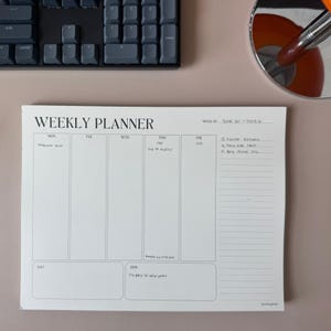 May include: A white weekly planner with the text "WEEKLY PLANNER" at the top. The planner has sections for each day of the week, with handwritten notes. A black keyboard and a mirror are in the background.