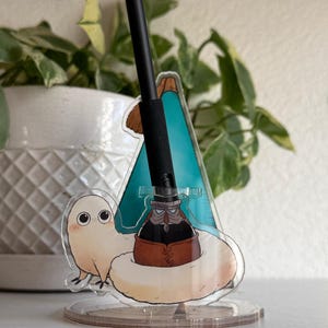 May include: A black pen rests in a clear acrylic stand featuring a cartoon character with a brown ink bottle. The stand has a clear base and a colorful design with a teal and brown background. The pen is black with a textured grip.