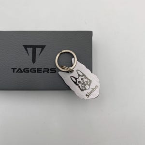 Puede incluir: Un llavero de metal plateado con un diseño de perro pastor alemán y el nombre "Simba". El llavero está sujeto a un anillo plateado y descansa sobre una caja gris con la palabra "TAGGERS" impresa.