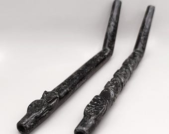 May include: Two black, carved stone pipes with a dragon head design. The pipes are angled with a curved shape and a hollowed-out opening. The stone has a marbled texture.