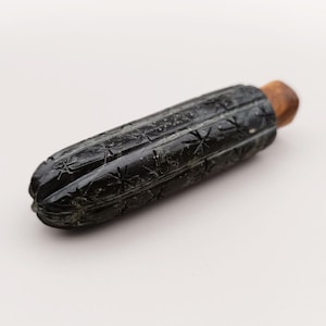 May include: A black bottle stopper with a carved, textured design and a wooden top. The stopper has a cylindrical shape with a series of star-like patterns carved into the surface. The wooden top is a light brown color.