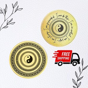May include: Two gold-coloured round metal discs with a black yin yang symbol in the centre. The discs have a black border with a repeating pattern. The top disc has the text "I am grounded, I can do this. I am strong, I am in control." The bottom disc has no text.