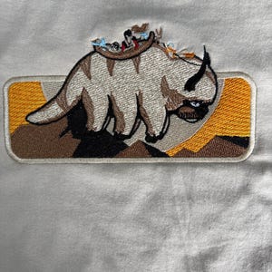 May include: Embroidered patch featuring a stylized Appa from Avatar: The Last Airbender. The patch shows Appa with riders, set against a landscape of mountains and a rectangular border with orange and brown accents. The patch is on a light-colored fabric.