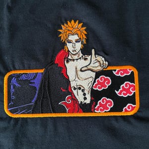 May include: Black t-shirt featuring an embroidered patch of an anime character with orange hair and a stern expression. The patch includes red clouds and a black background with a yellow border. The character is in a dynamic pose.