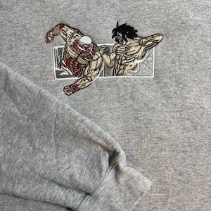May include: Gray hoodie featuring an embroidered design of two muscular figures in combat. The figures are detailed with tan, red, and black thread, set within a white rectangular frame. The hoodie has a drawstring hood and a ribbed cuff.