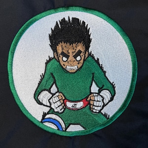 May include: Embroidered patch featuring a cartoon character in a green jumpsuit, white wrist wraps, and a red belt with a symbol. The character has spiky black hair and an intense expression, set against a white background with a green border.