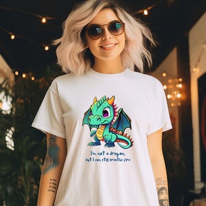 Pode incluir: Camiseta branca com um dragão de desenho animado em tons de verde, azul e amarelo. O dragão tem olhos roxos grandes e asas azuis. O texto abaixo diz: "Eu não sou um dragão, mas ainda posso cuspir fogo."