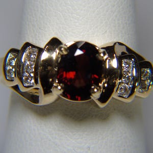 May include: A gold ring featuring a large, oval, deep red gemstone as the centerpiece. The ring has a decorative design with small, clear accent stones on each side of the central stone. The ring is set on a white surface.