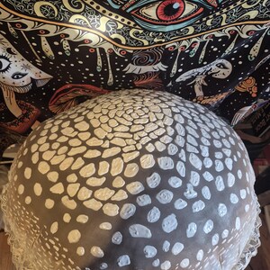 May include: A large, round decorative cushion with a dark grey base and white, irregular dot pattern. The cushion has a white lace trim around the edge. The background features a black tapestry with colourful psychedelic art.