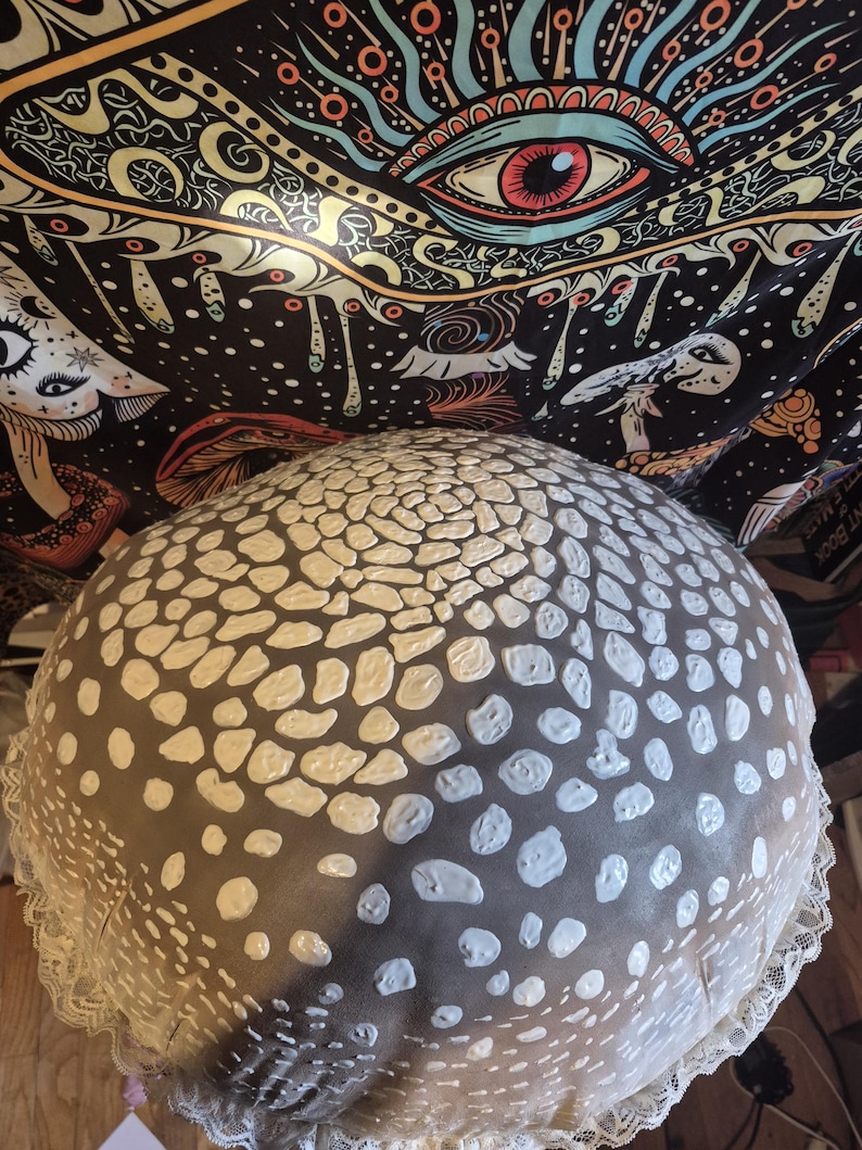May include: A large, round decorative cushion with a dark grey base and white, irregular dot pattern. The cushion has a white lace trim around the edge. The background features a black tapestry with colourful psychedelic art.