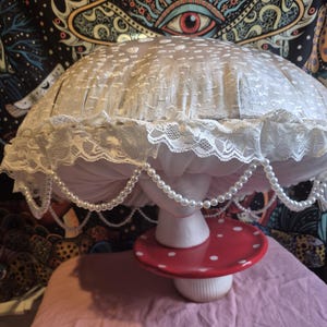 May include: A mushroom-shaped lamp with a white lampshade, lace trim, and a pearl strand. The red base features white spots. The lamp is set on a pink surface. The lamp is a decorative item.
